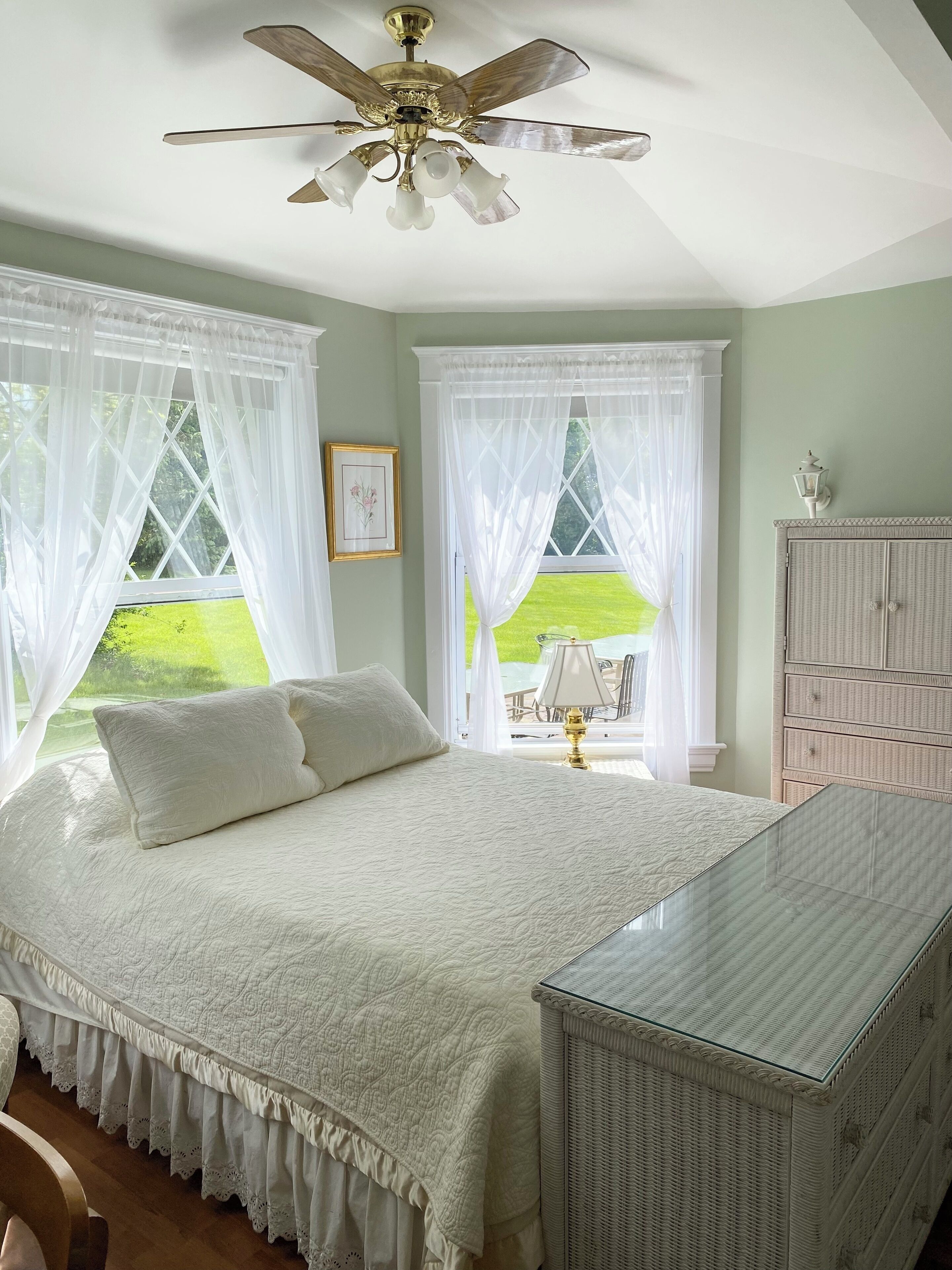 A bedroom with a bed, a glass-top dresser, a ceiling fan, and two windows with white curtains.