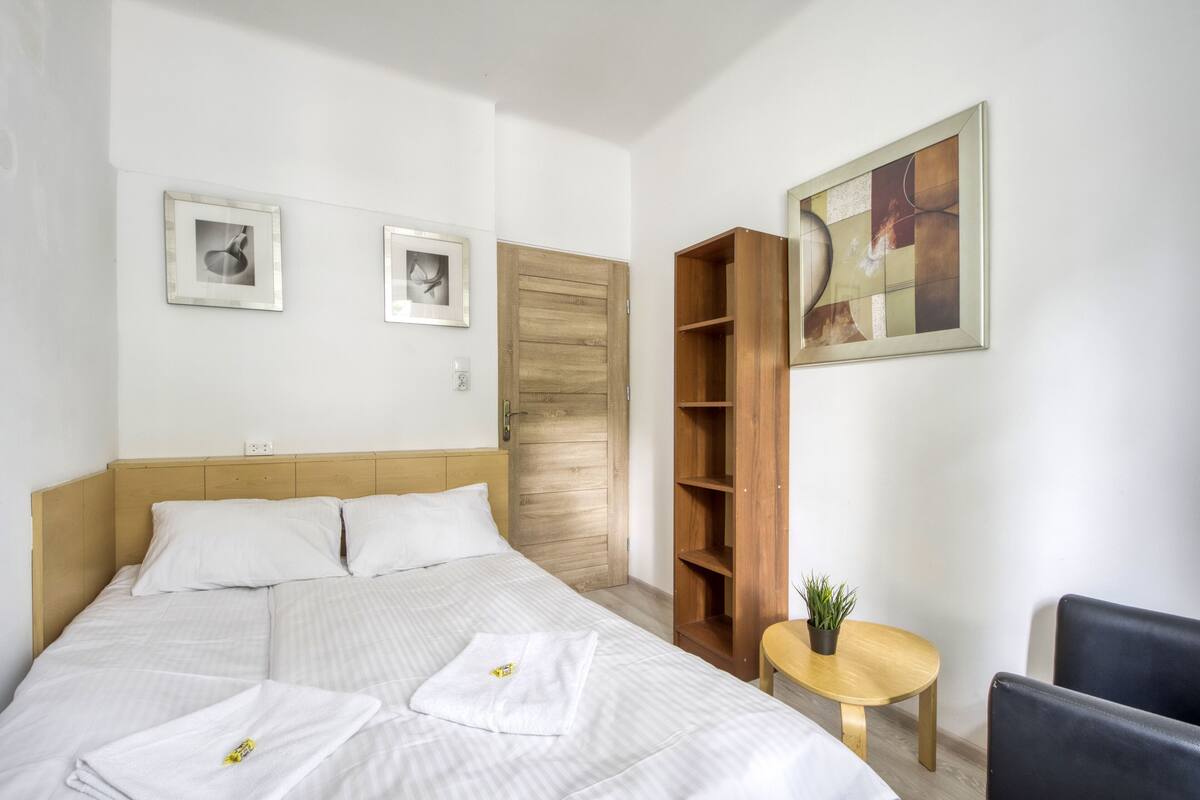 Ein Hotelzimmer mit einem Bett, zwei gerahmten Bildern, einer Holztür, einem Holzregal, einem kleinen runden Tisch mit einer Pflanze und zwei Stühlen.