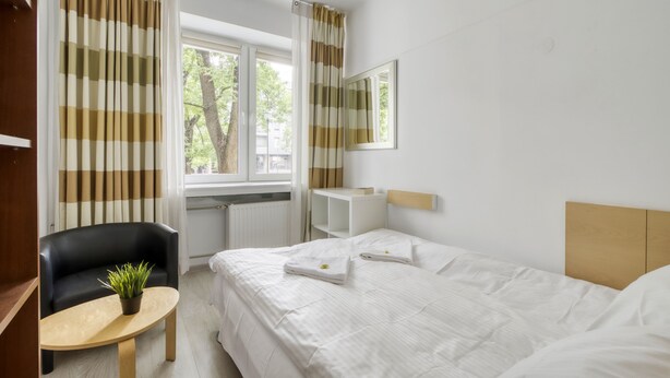 Ein Hotelzimmer mit Bett, Sessel, kleinem Tisch mit Pflanze, Fenster mit Vorhängen und Spiegel an der Wand.