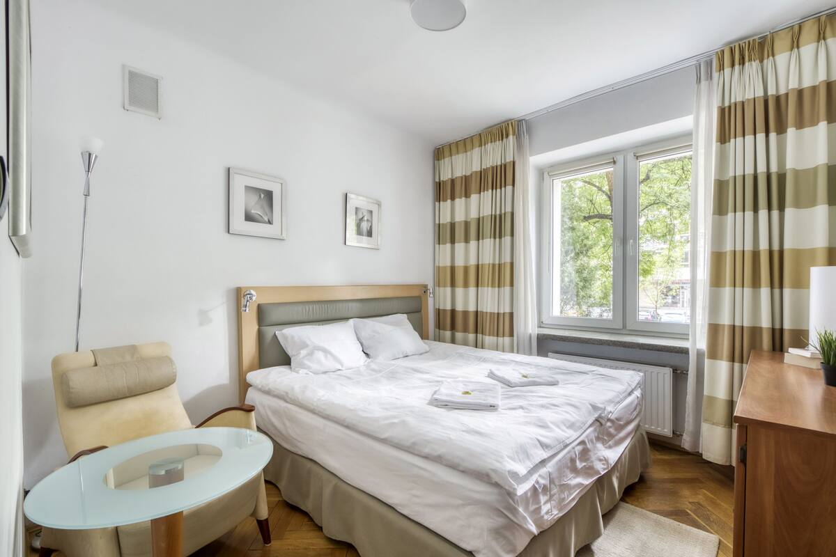 Ein Hotelzimmer mit Bett, Stuhl, kleinem Tisch und einem Fenster mit Vorhängen.