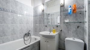Ein modernes Badezimmer mit Badewanne, Waschbecken und Toilette. Über dem Waschbecken befinden sich Glastage mit Pflegeprodukten.