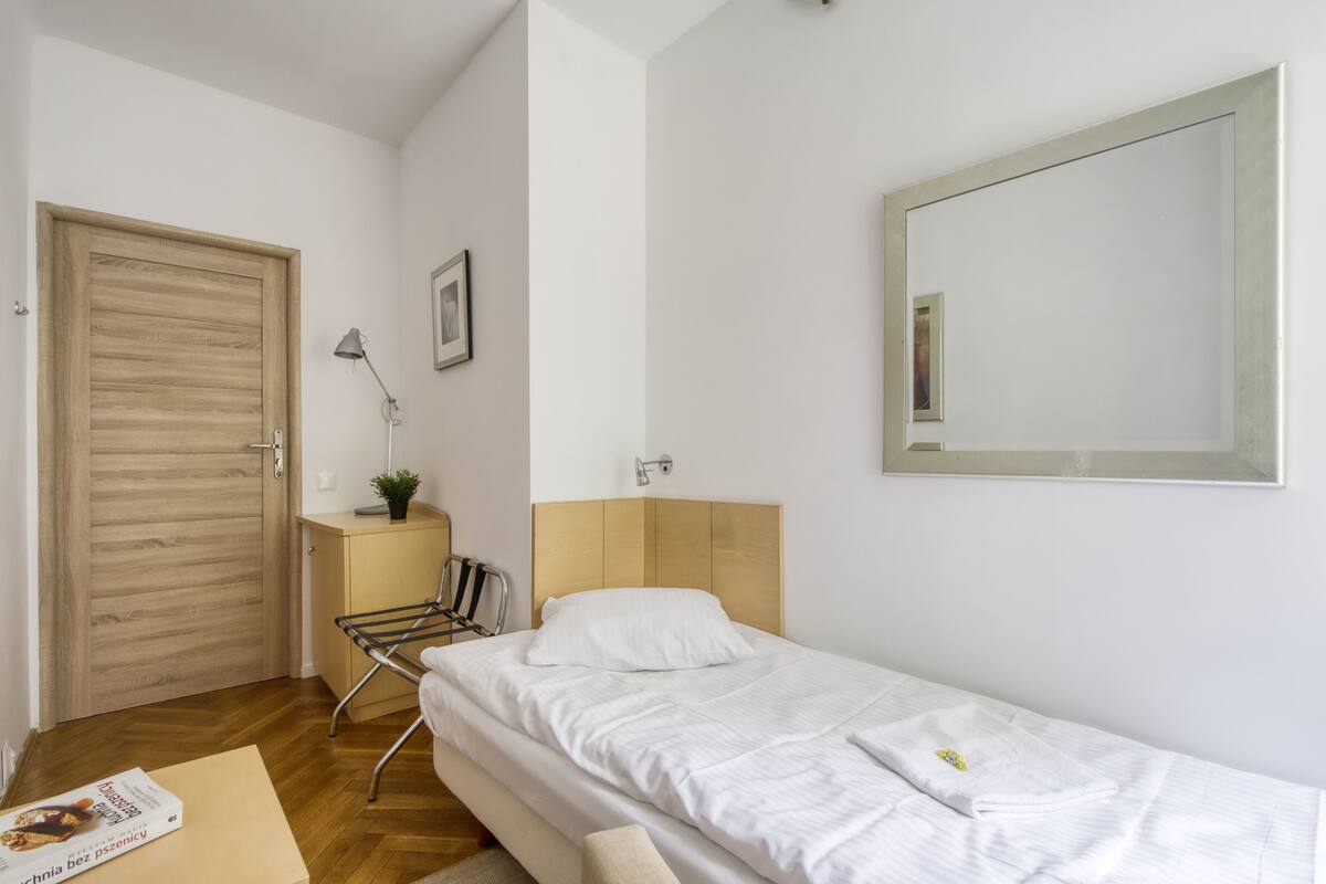 Ein Zimmer mit einem Einzelbett, einer Holztür, einem kleinen Nachttisch mit Lampe, einem Spiegel an der Wand und einem Stuhl.