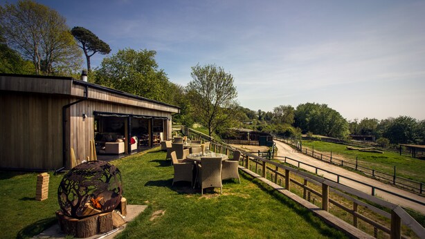 A rustic outdoor seating area with a round table and chairs, a fire pit, and a wooden deck. - Treehouse Hotel - At Port Lympne Reserve (Hythe)