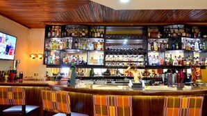A bar area with multiple shelves of liquor bottles, a bartender, and bar stools. - The Cape Hotel (Monrovia)