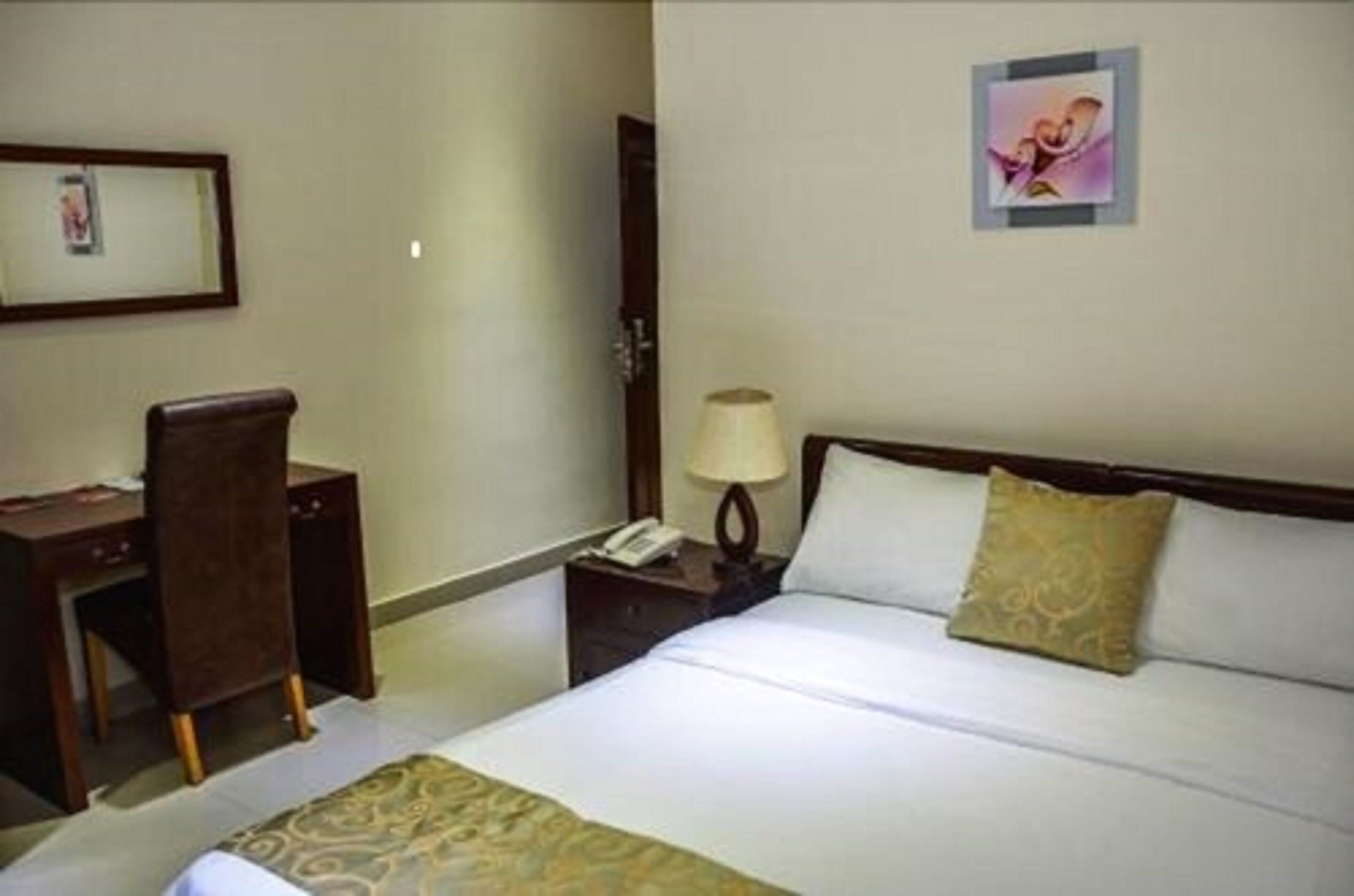 A hotel room with a bed, a desk, a chair, a nightstand with a lamp, and a framed picture on the wall.