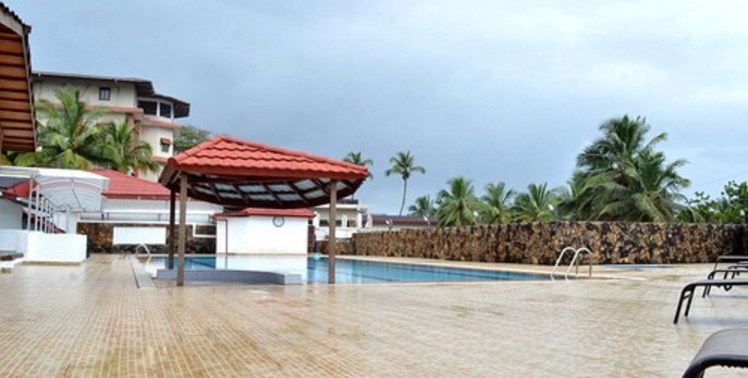 Zona de piscina con un área de estar techada, palmeras y un muro de piedra.