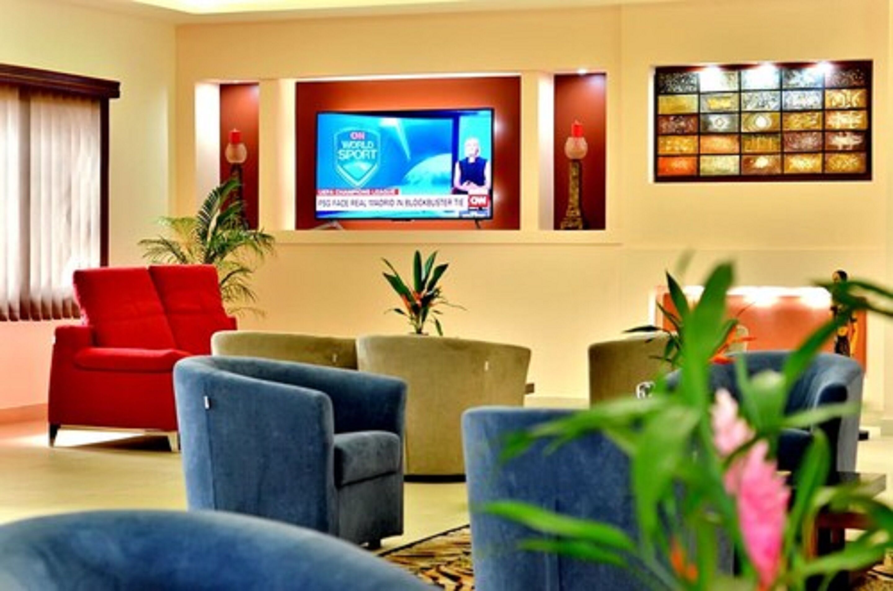 Um saguão de hotel com uma televisão exibindo um canal esportivo, assentos confortáveis e painéis decorativos na parede.