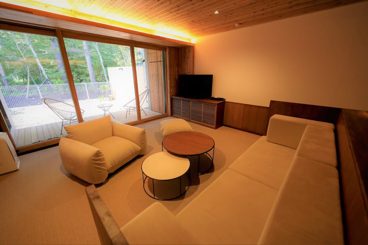 A modern living room with a sofa, two round tables, a TV, and a sliding glass door leading to an outdoor area with chairs.