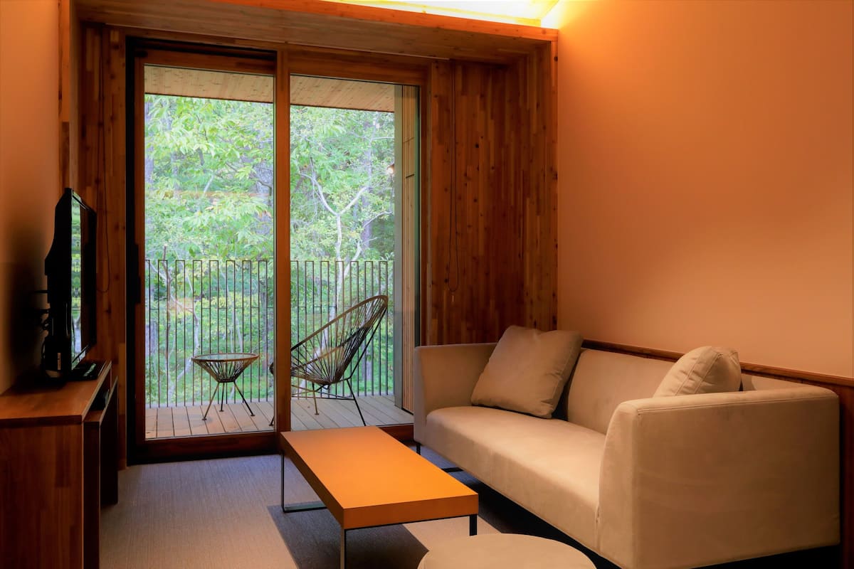 A cozy living room with a sofa, a coffee table, and a TV on a wooden stand. A chair is placed by the window, and a balcony with a railing is visible.