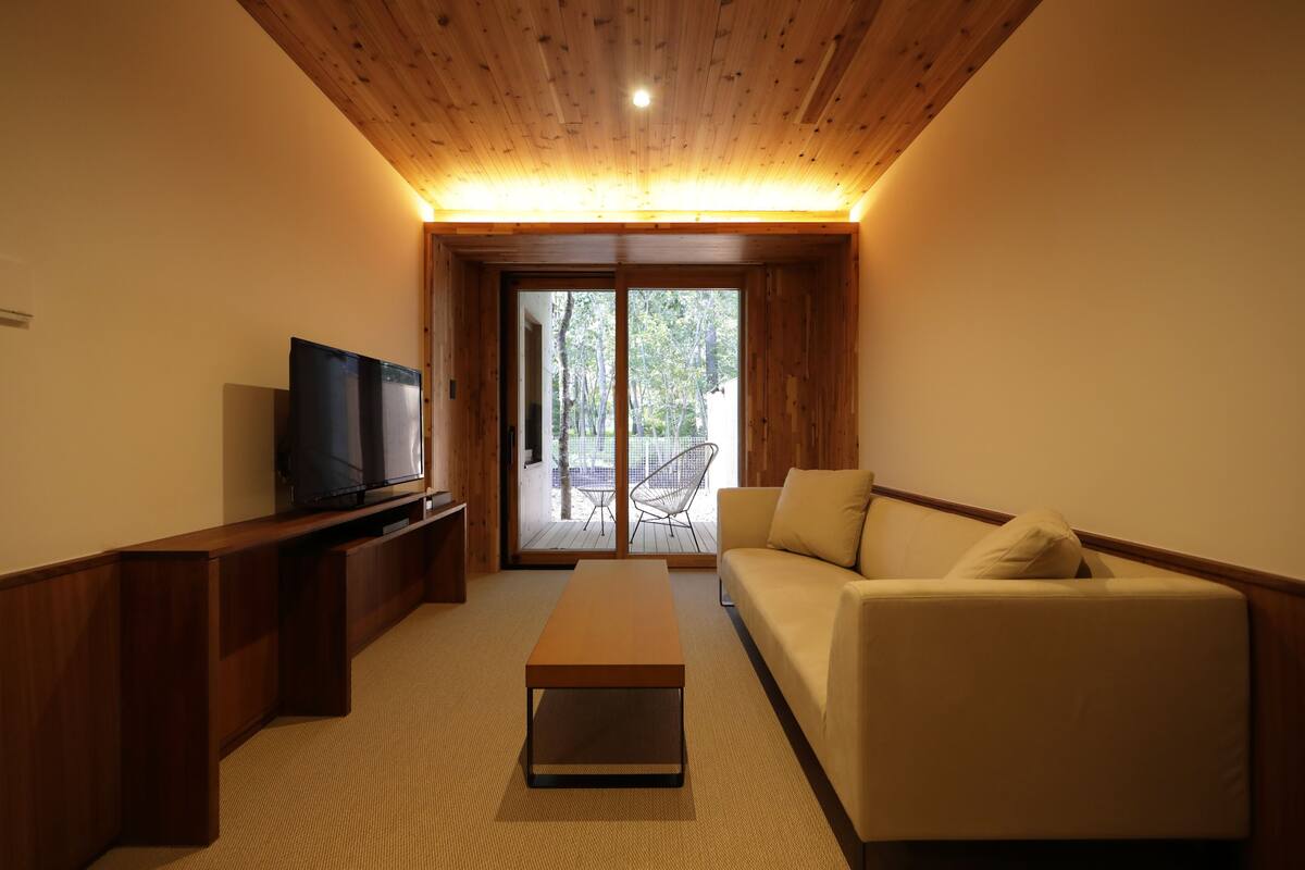 A modern living room with a wooden ceiling, a flat-screen TV on a wooden stand, a beige sofa, and a glass door leading to an outdoor area.