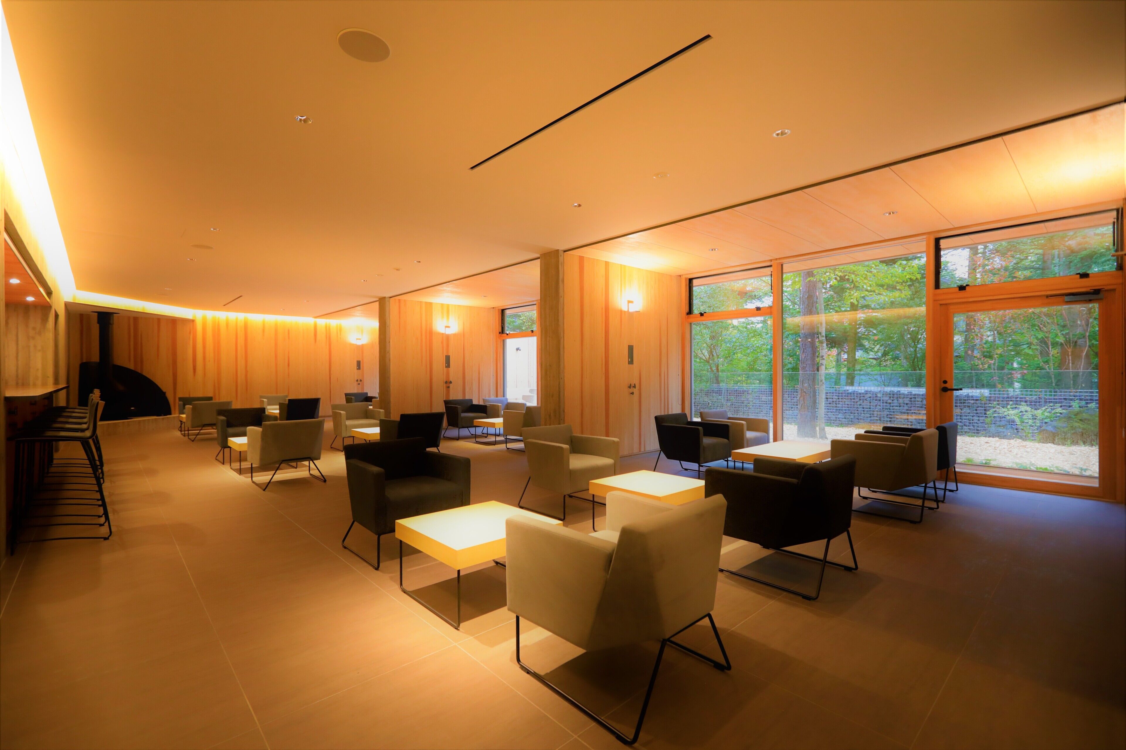 A modern interior with a mix of seating options, including sofas and chairs, arranged around tables. Large windows provide natural light and views of greenery outside.