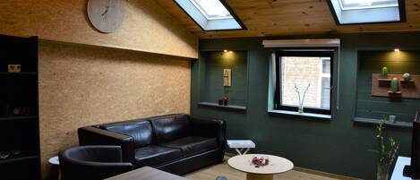 Un intérieur moderne avec un plafond en bois, des puits de lumière, un canapé en cuir noir, une table ronde et une horloge au mur.
