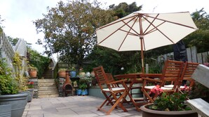 Eine Terrasse im Freien mit einem Holztisch, Stühlen, einem großen Sonnenschirm und verschiedenen Blumentöpfen.