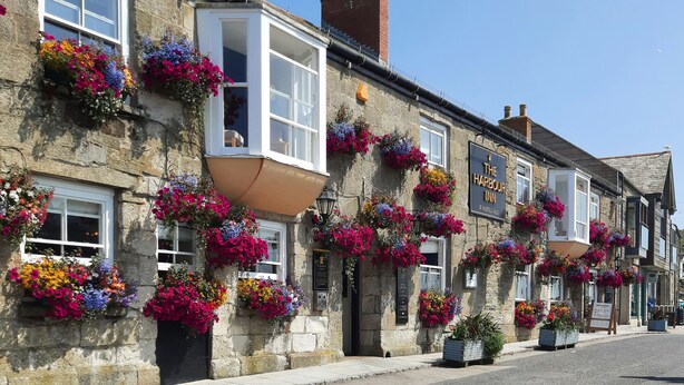 Ein traditionelles Steinhaus mit zahlreichen Fenstern, von denen einige mit Blumenkästen versehen sind. Am Gebäude ist ein Schild mit der Aufschrift "The Harbour Inn" angebracht.