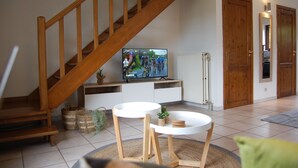 Un salon avec un escalier en bois, un téléviseur diffusant une course cycliste, une table ronde blanche et un coussin vert.