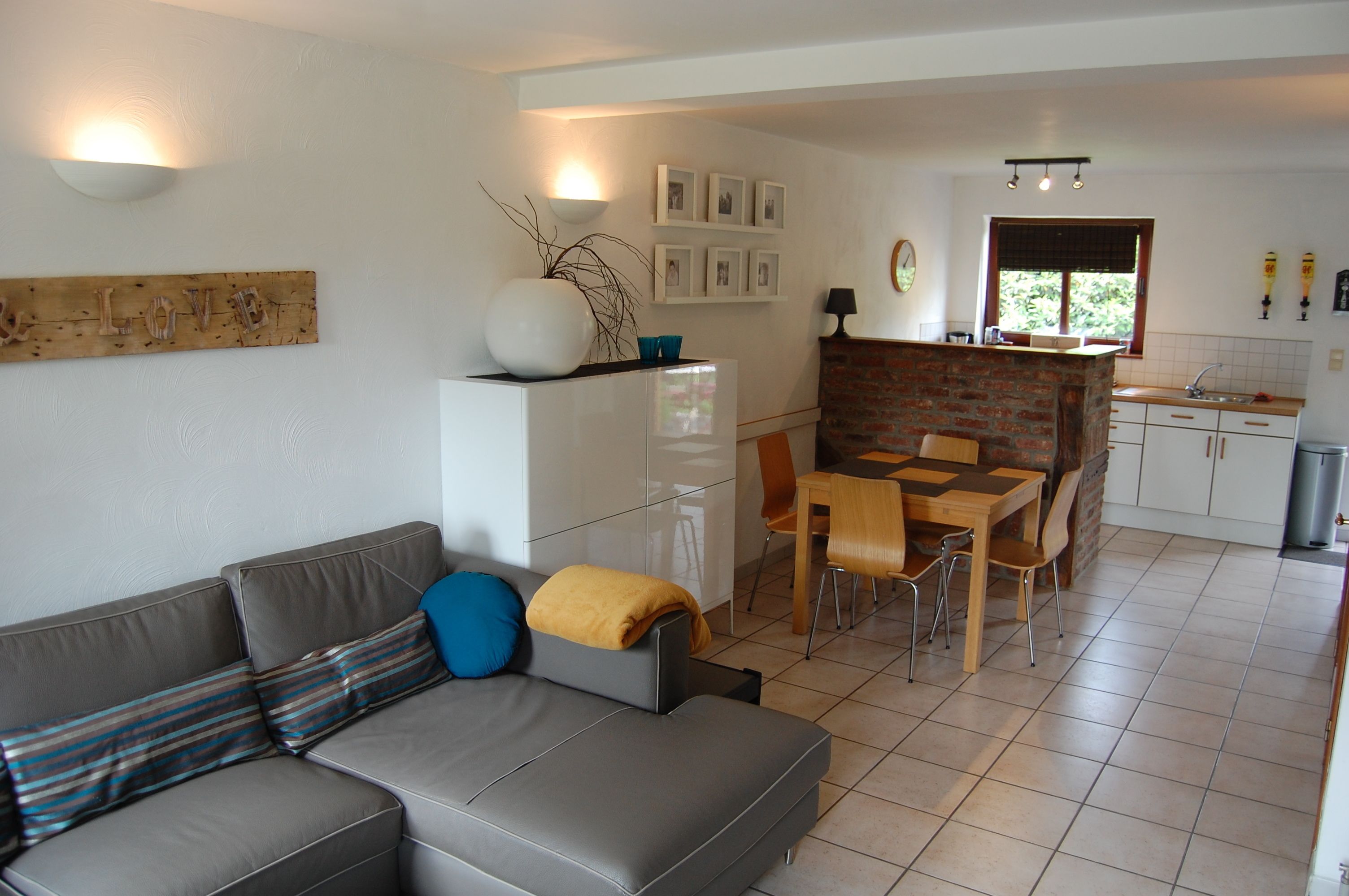 Een moderne woonkamer met een grijze bank, een eettafel met stoelen, een keuken met een bakstenen muur en een witte koelkast.