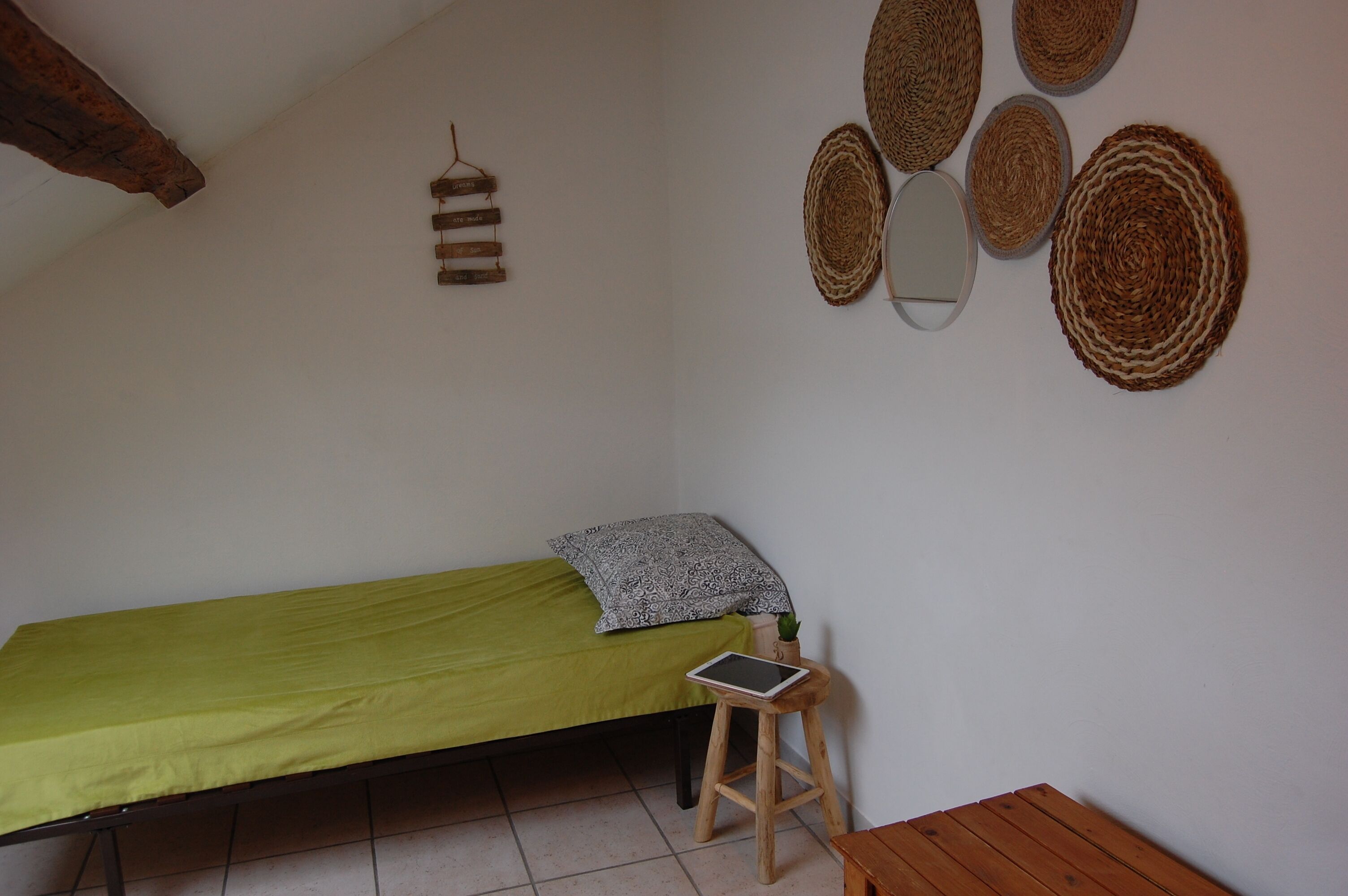 A small, single-bed room with a green bedspread, a wooden stool with a tablet, and woven wall decorations.