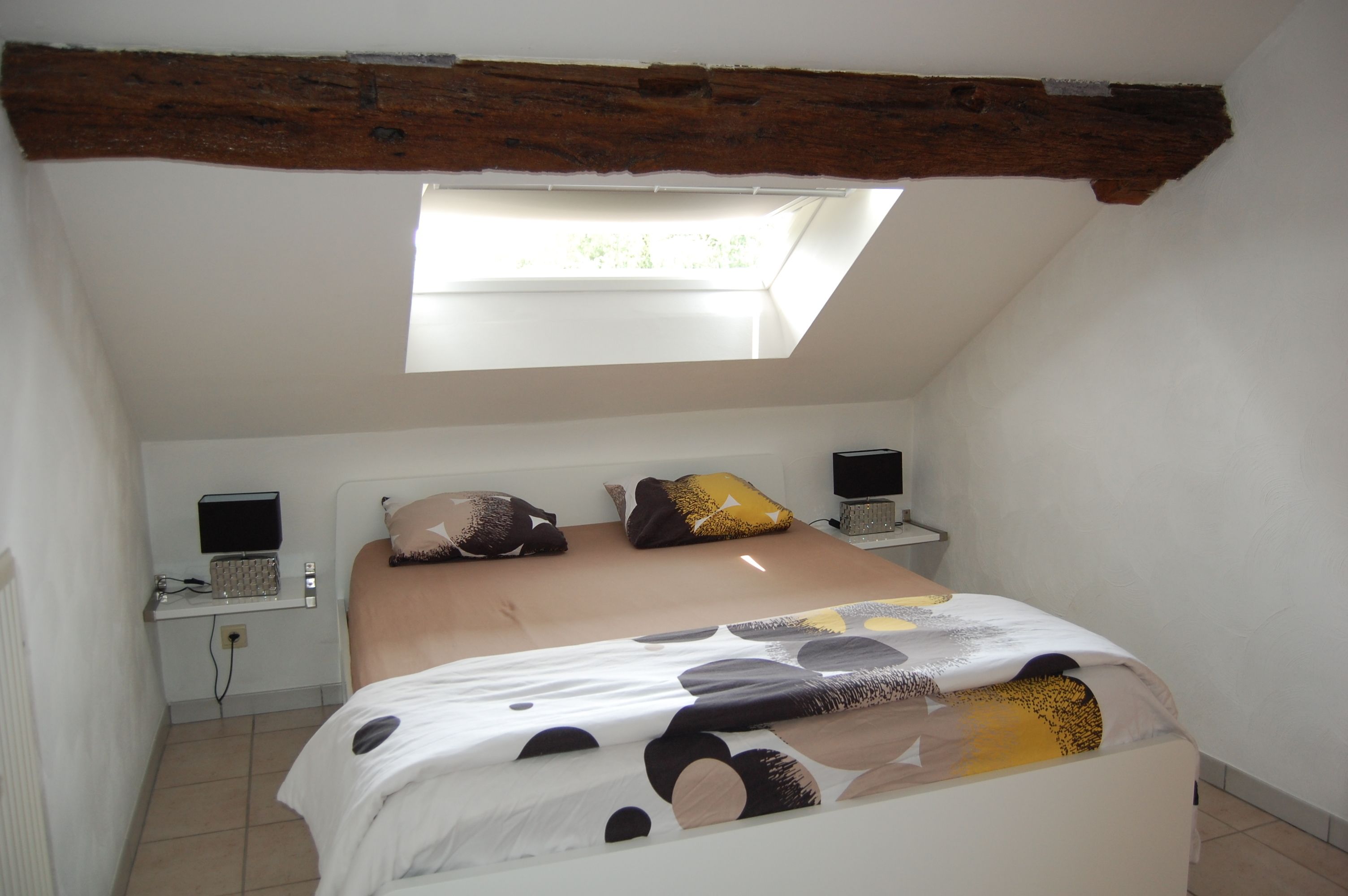 A bedroom with a sloped ceiling, a skylight, a bed with a patterned bedspread, two bedside tables with lamps, and a wall-mounted television.