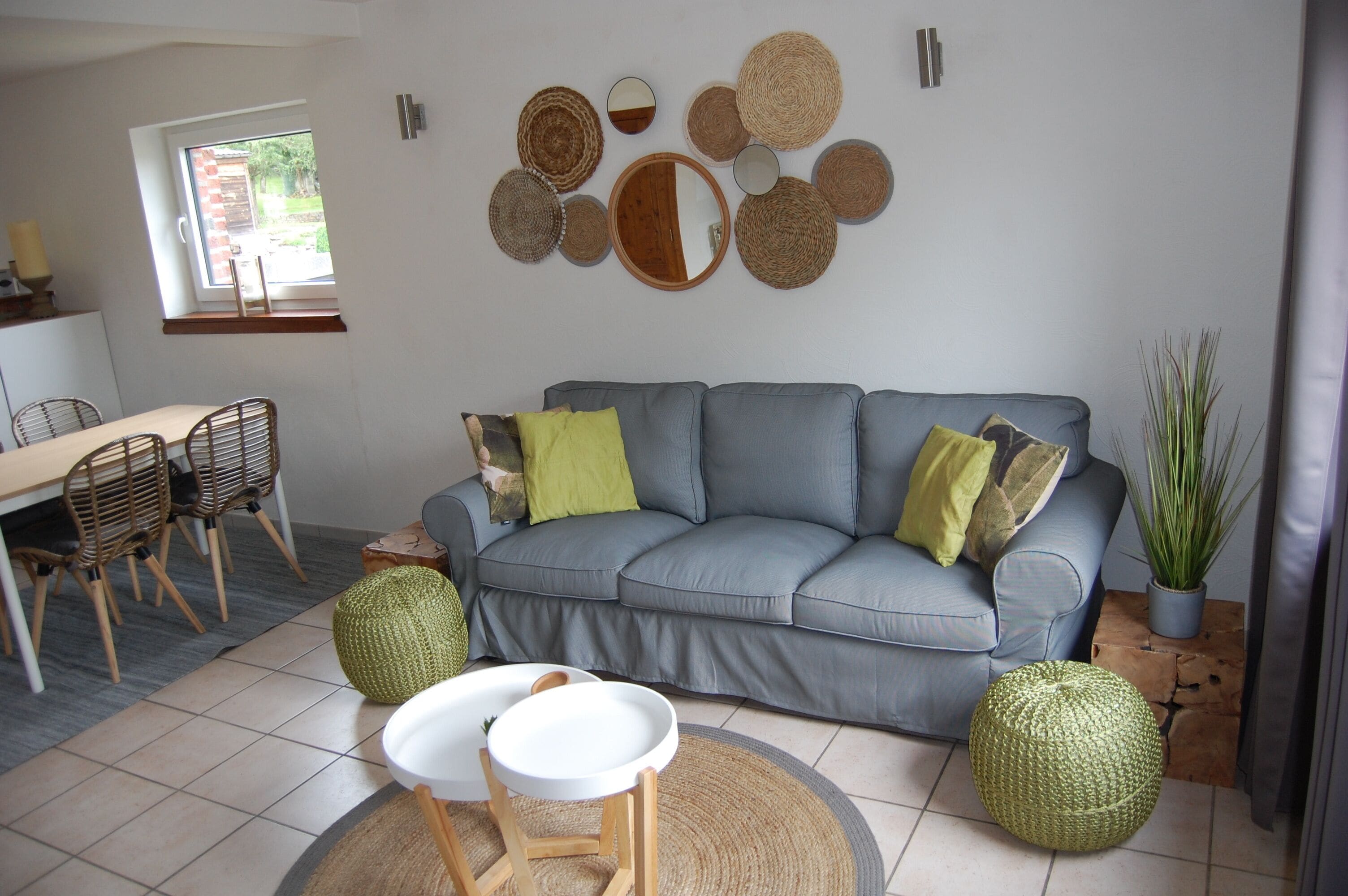 Sala de estar com sofá cinza, dois pufes redondos trançados, canto de jantar com mesa e cadeiras, e um espelho grande na parede.