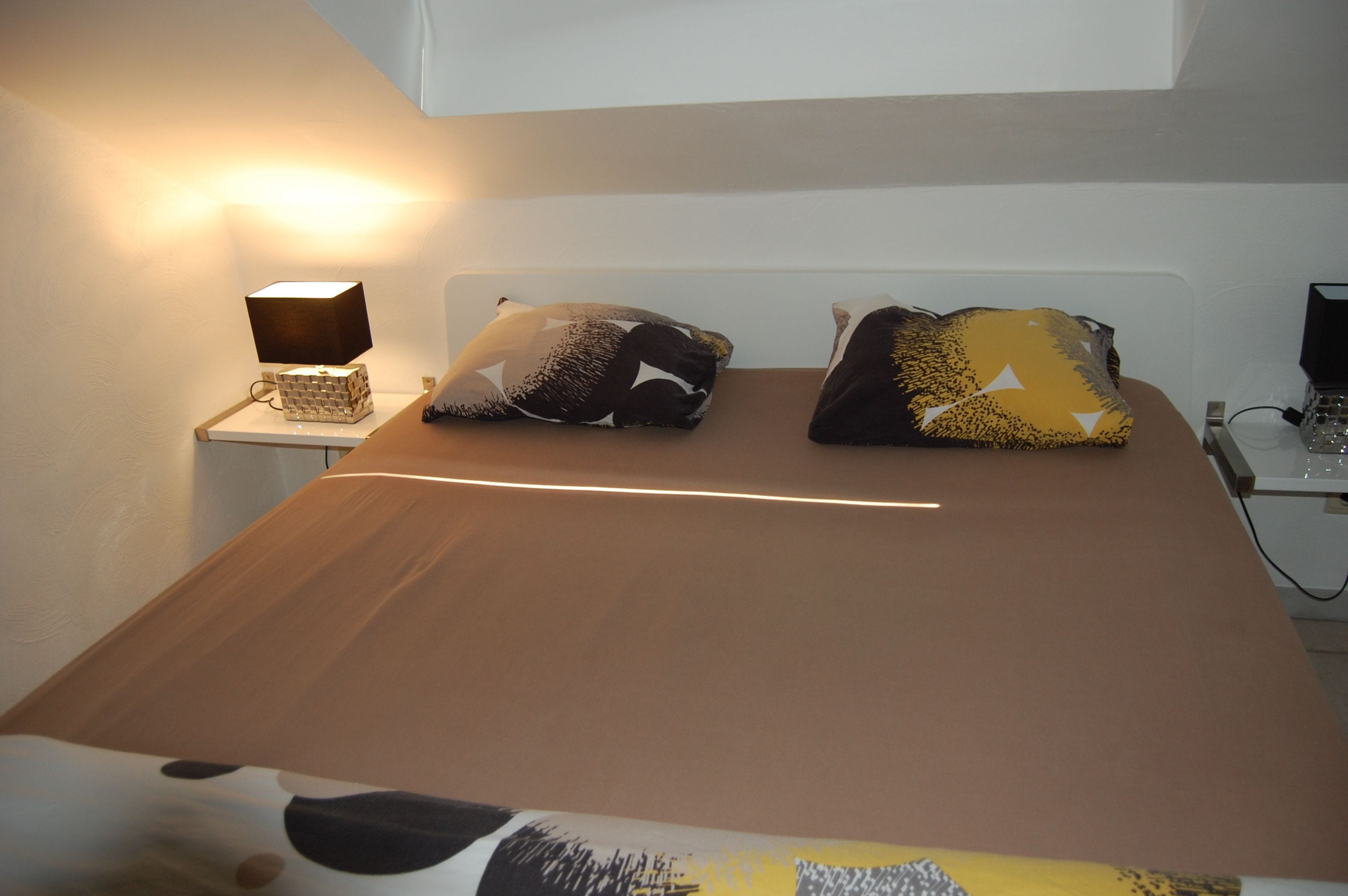 Een opgemaakt bed met een bruine sprei, twee gestreepte kussens, een bedlampje en een laptop op een bureau naast het bed.