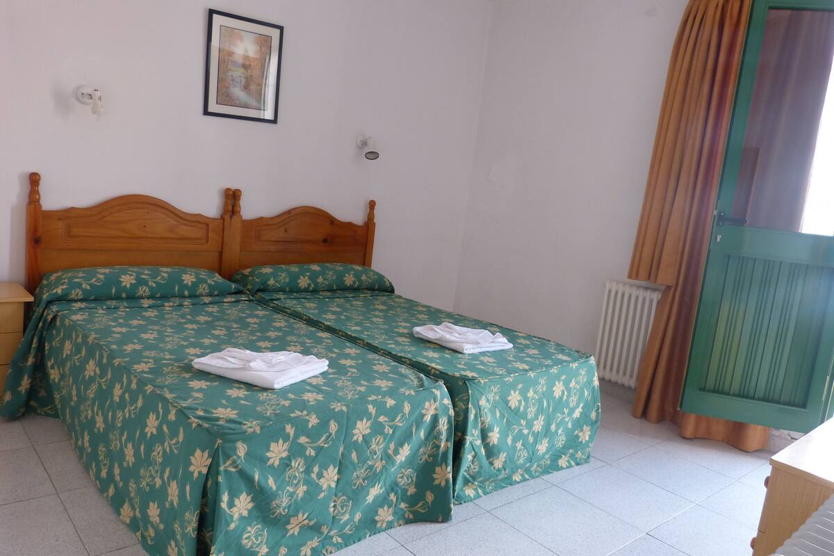Un dormitorio con cama de madera, ropa de cama floral verde, un cuadro enmarcado en la pared y una ventana con cortinas.