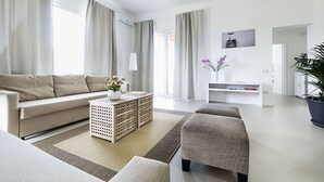 Een moderne woonkamer met een bank, een poef en een salontafel met bloemen. Er is een witte tv-meubel en een potplant.