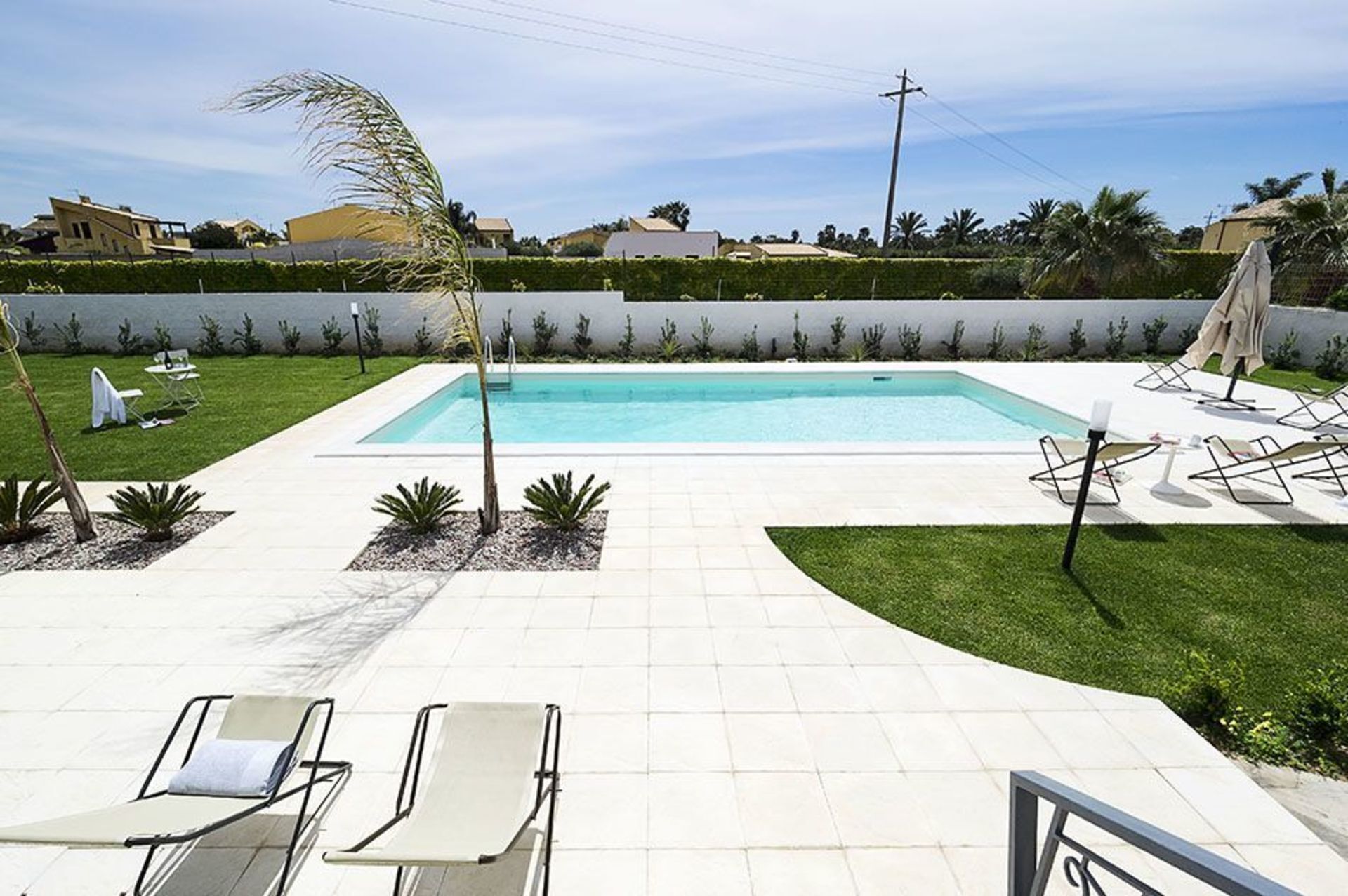 A rectangular swimming pool with a tiled deck, surrounded by a low hedge, and equipped with lounge chairs and a sun umbrella.