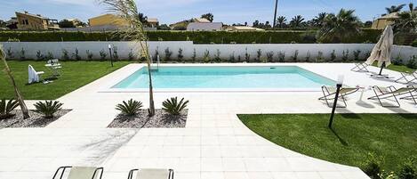 A rectangular swimming pool with a tiled deck, surrounded by a low hedge, and equipped with lounge chairs and a sun umbrella.