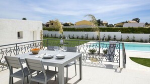 Een terras met een eettafel, een zwembad op de achtergrond en een wit gebouw met een klein raam.