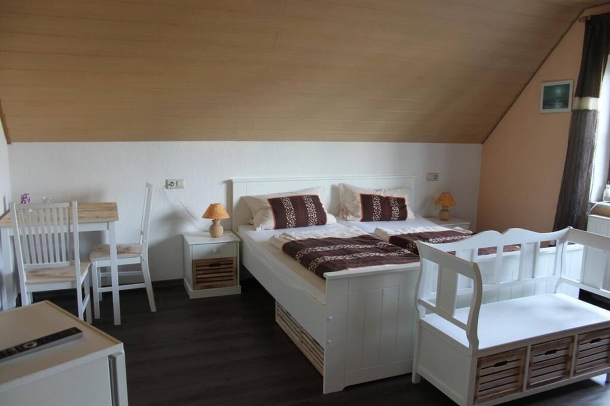 Une chambre individuelle avec un plafond en bois, un lit avec une literie blanche et des coussins à motifs, une table de chevet avec une lampe et une chaise.