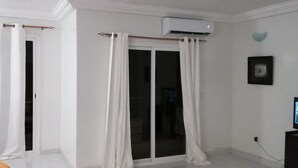 Een kamer met witte wanden, een schuifdeur met glas, witte gordijnen, een airconditioning en een klein zwart tv-meubel, plus één oranje kussen.
