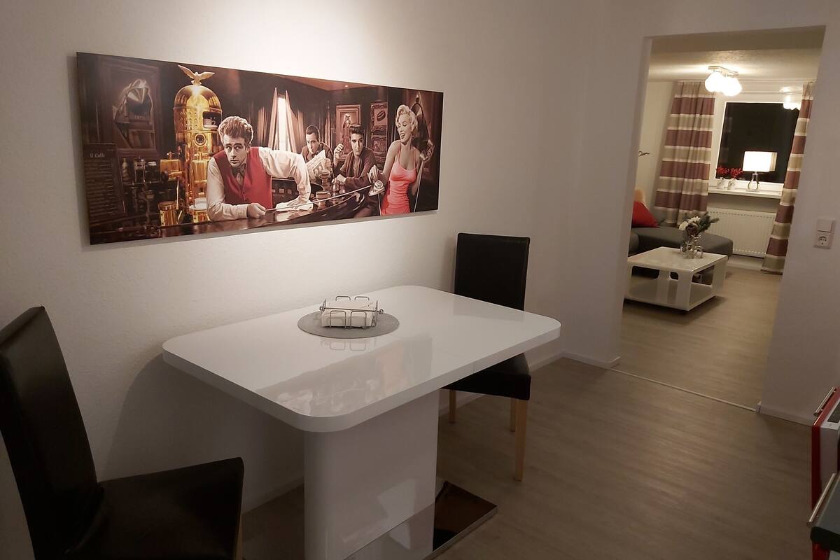 Un coin repas avec une table blanche, des chaises noires et un tableau au mur. On aperçoit une petite table avec un vase au fond.