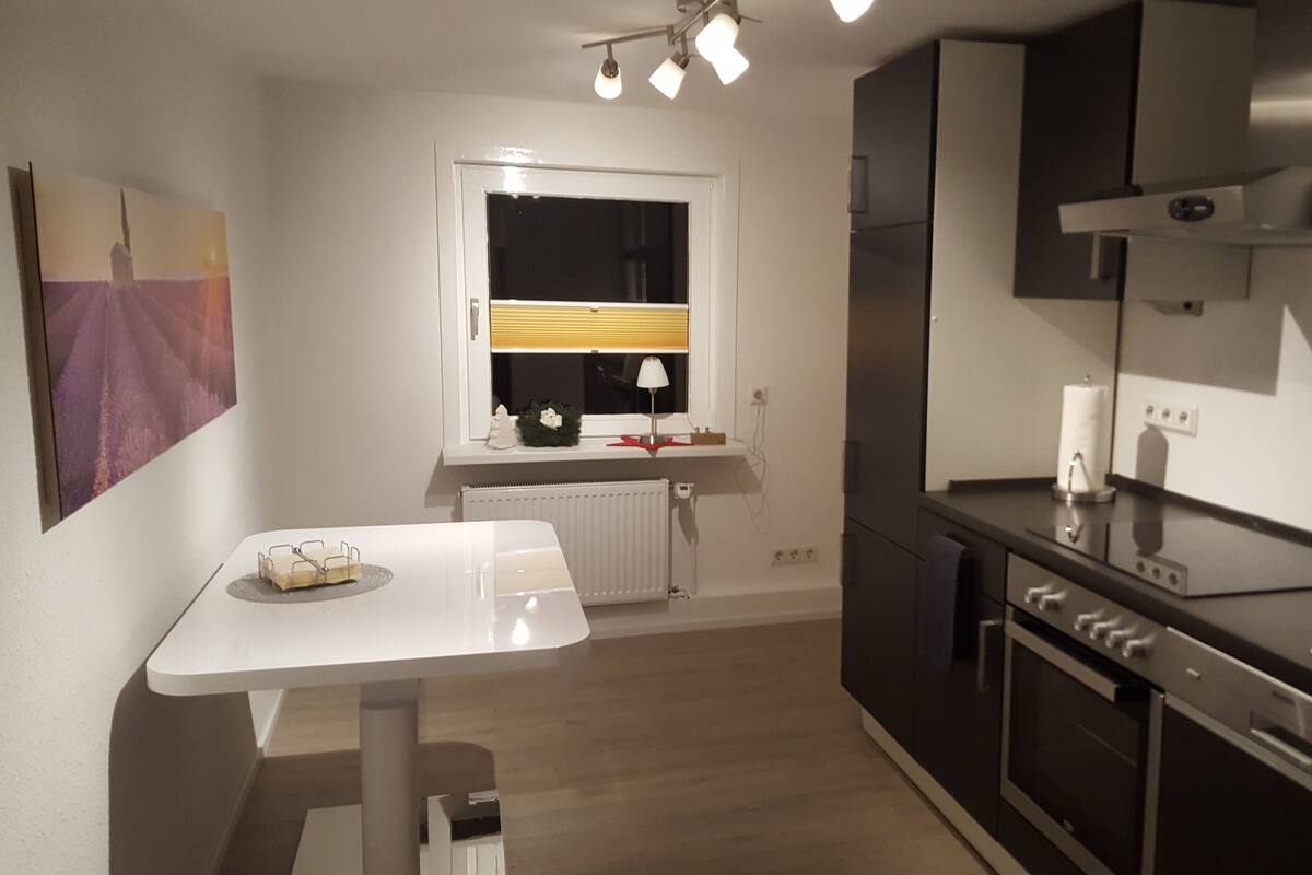 Une cuisine moderne avec une table blanche, une fenêtre avec des stores, un radiateur et des appareils électroménagers intégrés.