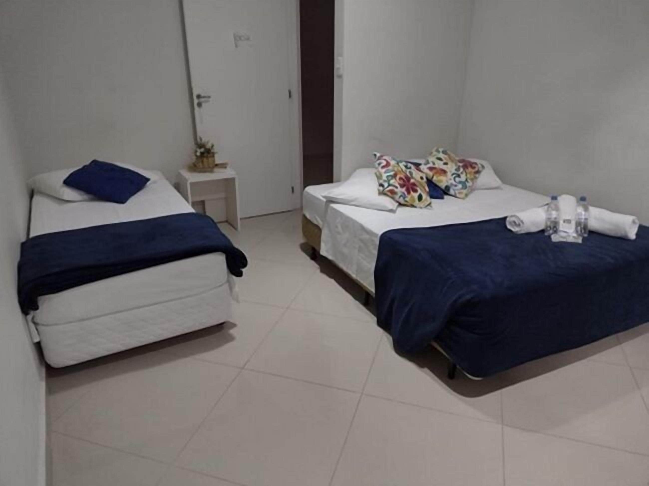 Een hotelkamer met twee bedden, elk met een blauwe deken en kussens. Er staat een klein nachtkastje met een plant en een fles water.