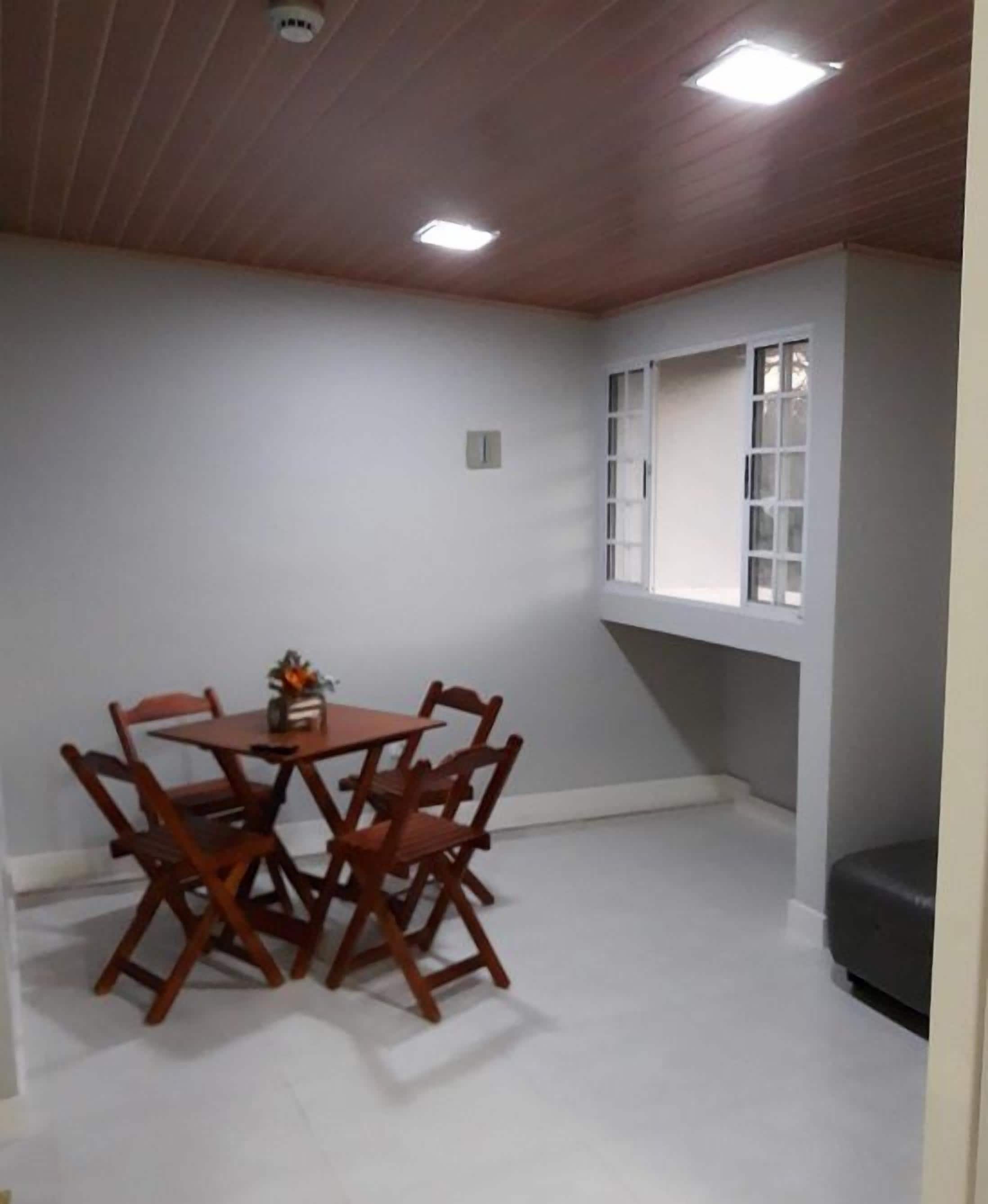 Quarto com mesa de jantar e cadeiras, janela com persianas e iluminação embutida no teto.