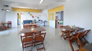 A spacious dining area with multiple wooden tables and chairs, a kitchen area in the background, and a yellow accent wall. - Pousada Top (Mairipora)