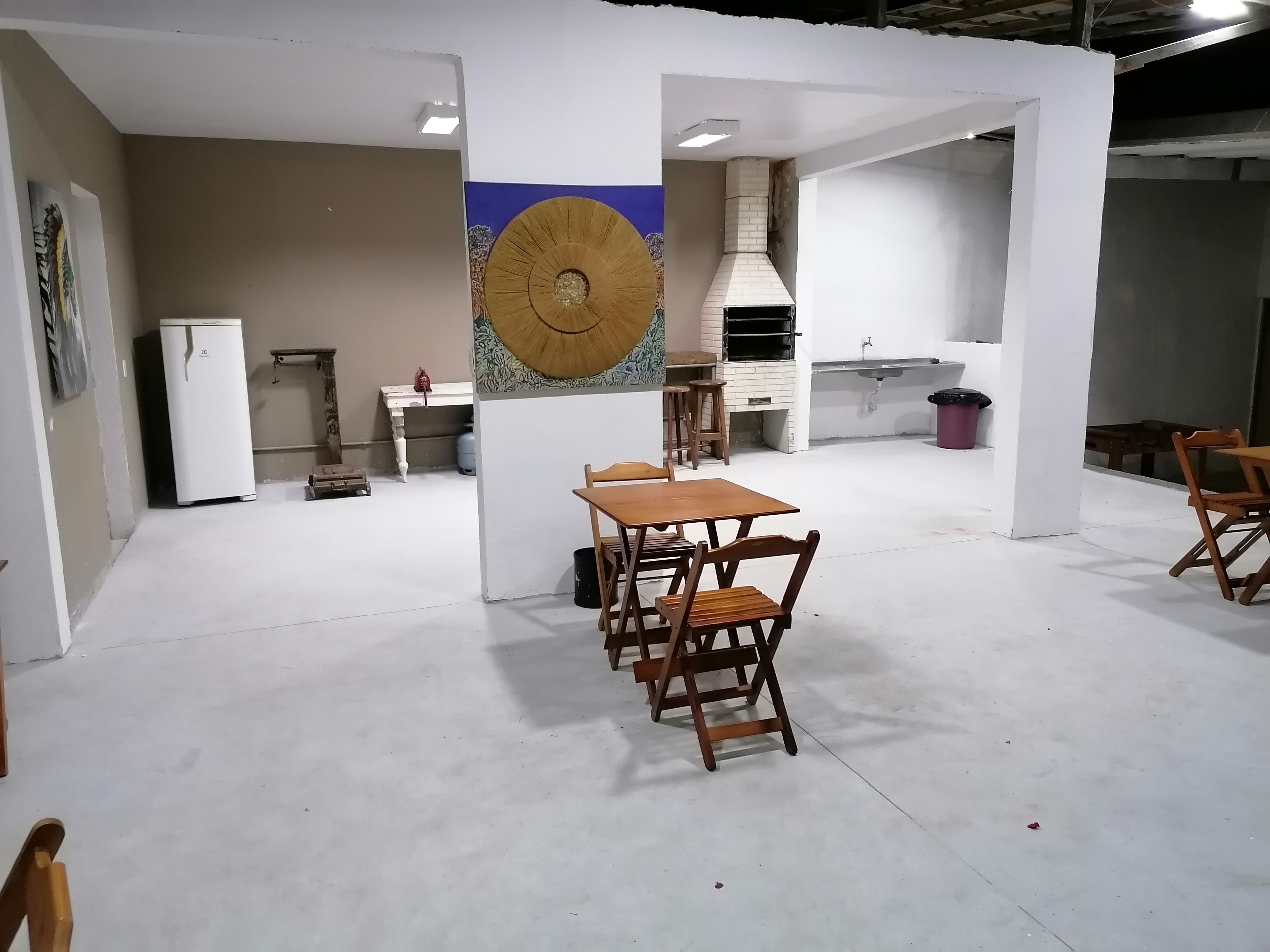 Una habitación amplia con comedor que incluye una mesa y sillas de madera, un refrigerador blanco, zona de cocina con estufa y lavaplatos, y un elemento decorativo en la pared.