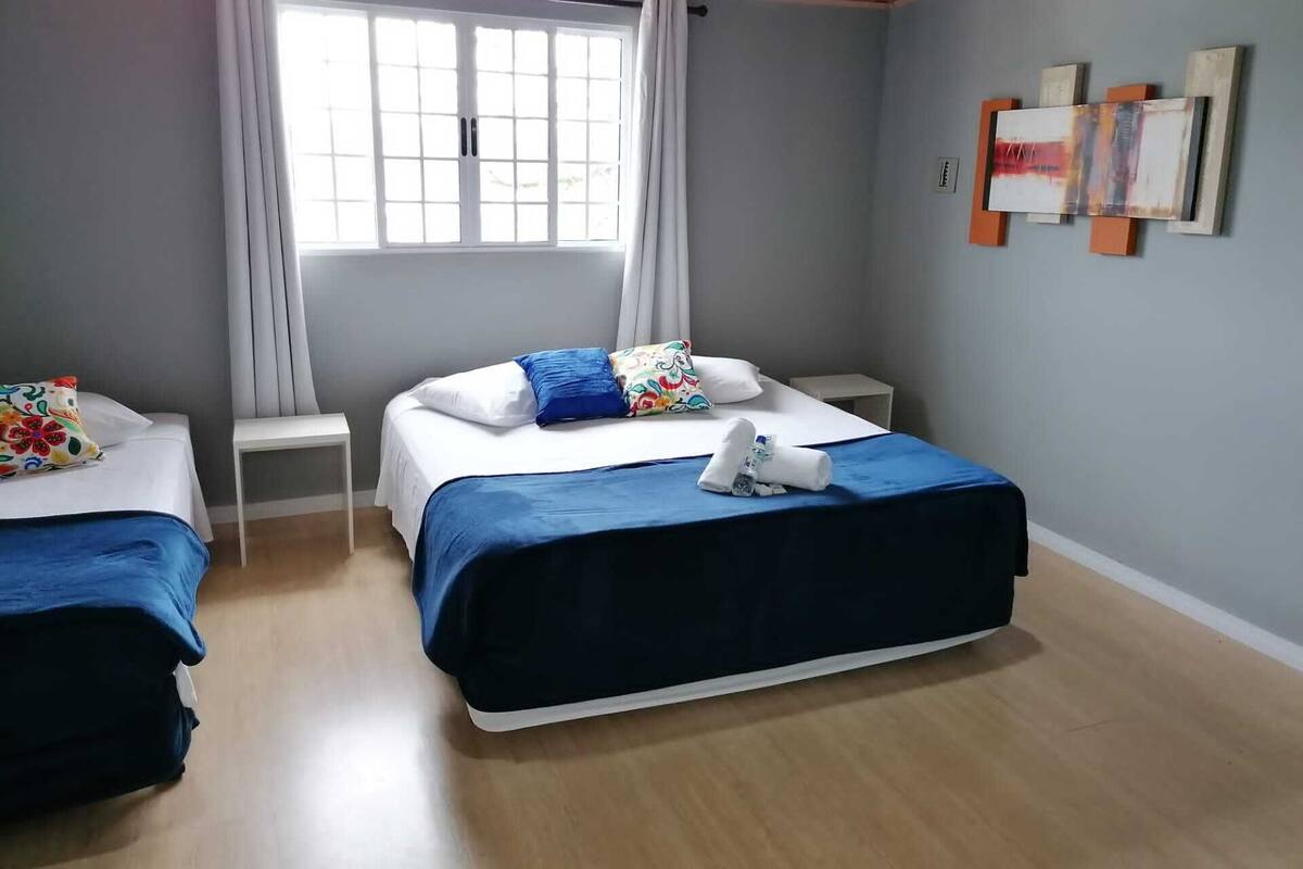 A hotel room with two beds, a window with curtains, a small bedside table, and artwork on the wall.