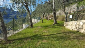 A grassy pathway flanked by trees, leading to a stone wall and a fenced area with a view of a lake and mountains. - Agricampeggio Agri-camping Relax (Brenzone sul Garda)