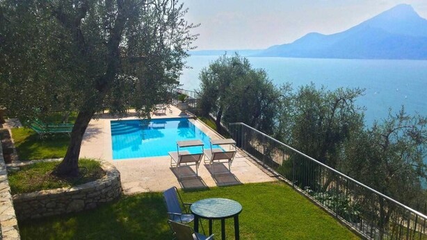 A swimming pool with lounge chairs and a table, surrounded by a stone border and grass, with a view of a lake and mountains in the background. - Agricampeggio Agri-camping Relax (Brenzone sul Garda)