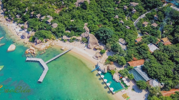 An aerial view of a coastal resort with a beach, clear waters, a wooden pier, and thatched-roof buildings. - Sao Bien Cam Ranh (Nam Cam Ranh)