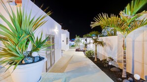 Un étroit sentier bordé de murs blancs et de plantes en pot, menant à l’entrée d’un bâtiment.