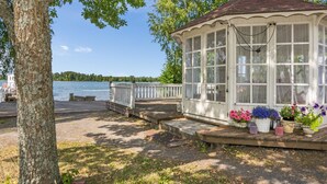 Um gazebo de madeira com estrutura de vidro, com vista para um lago, rodeado por árvores e um píer.