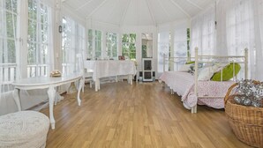 Quarto com piso de madeira, mobiliário branco, cama com roupa de cama floral e janelas amplas.