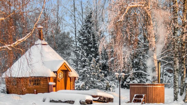 Uma cabana de madeira com telhado coberto de neve, uma chaminé soltando fumaça e um pequeno galpão de madeira por perto.