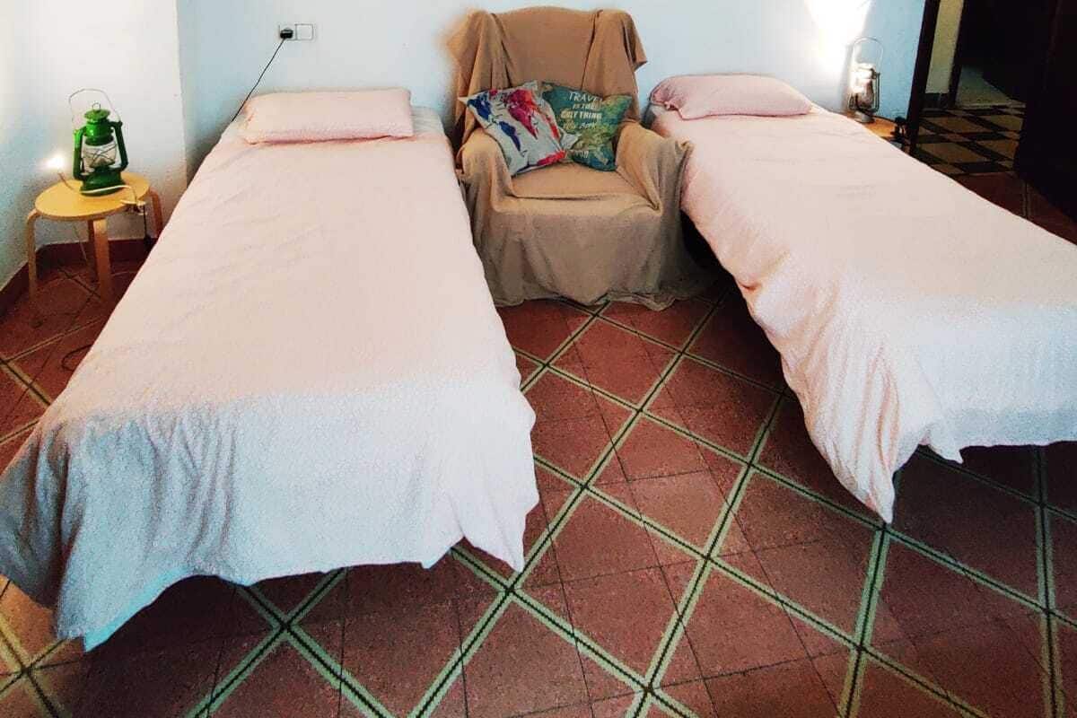 A small, simple room with two single beds, a chair with a colorful cushion, a small table with a green lantern, and a tiled floor.