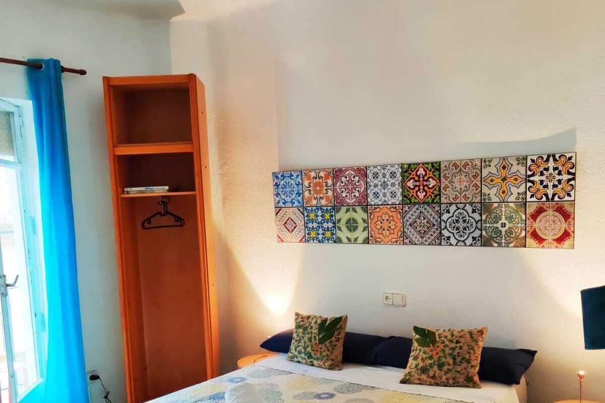 A bedroom with a bed, a wall-mounted tile art piece, a wooden wardrobe, and a blue curtain.