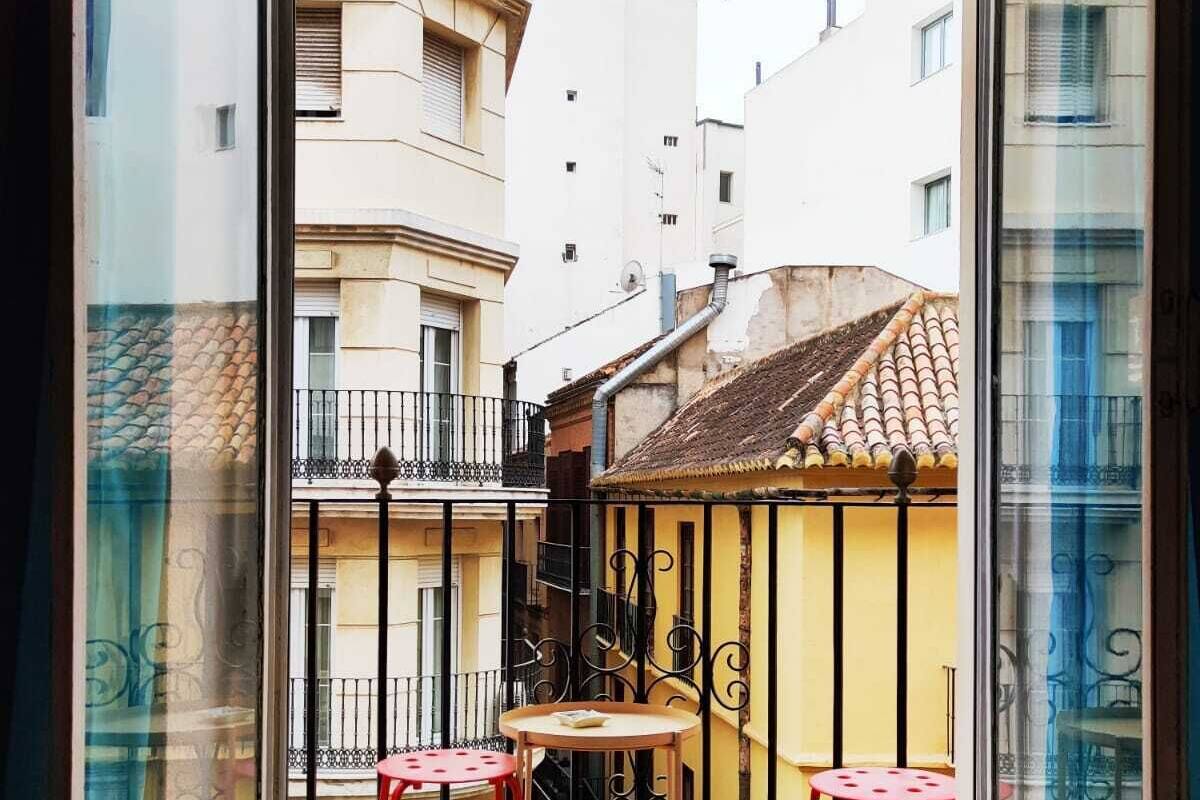 An open balcony with two red chairs and a small table, overlooking a narrow street with buildings.