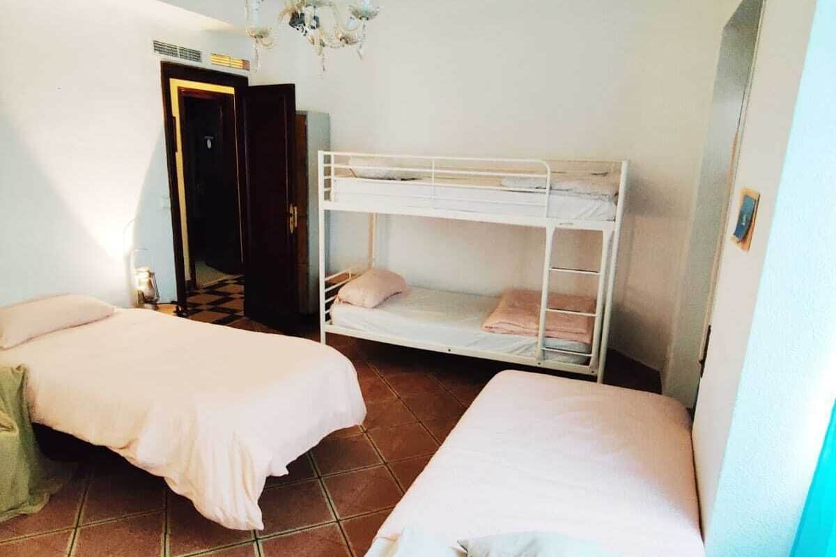 A dormitory room with two single beds, a bunk bed, a chandelier, and a door leading to another room.