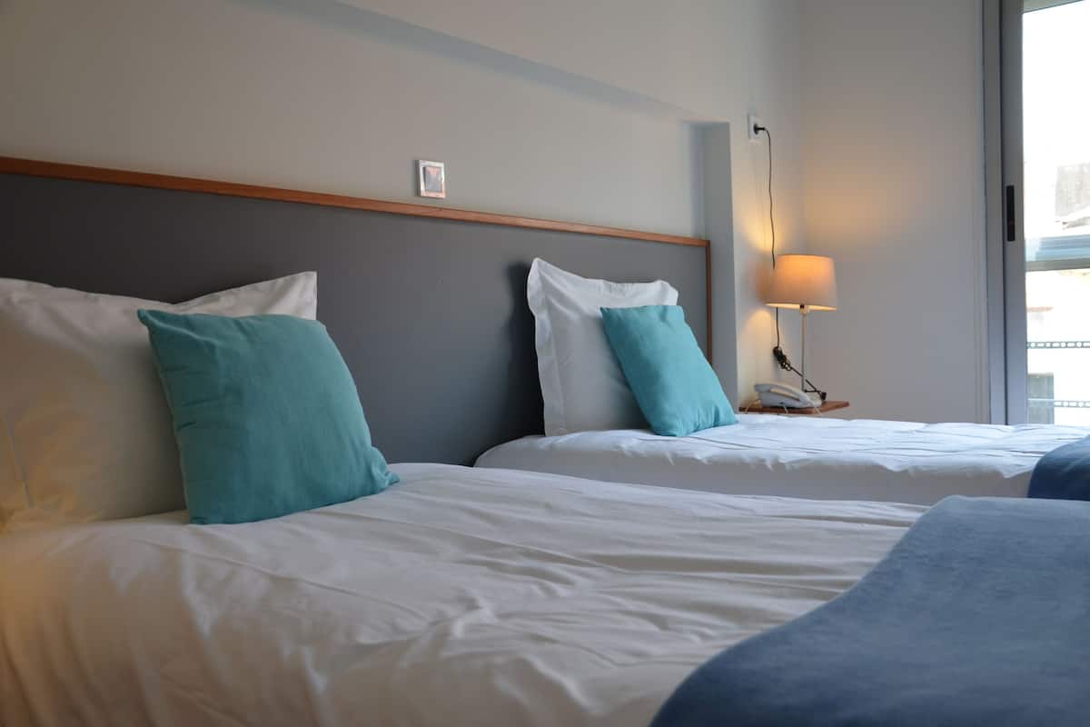 A hotel room with two beds, each with white bedding and teal pillows. There is a bedside table with a lamp and a phone.